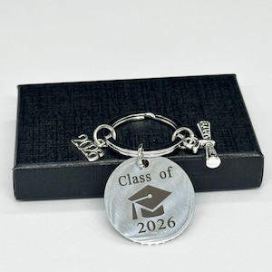 Könnte beinhalten: Silberner Abschluss-Schlüsselanhänger mit einem runden Anhänger mit der Gravur "Class of 2026" und einer Abschlusskappe. Enthält Charms mit dem Jahr "2026" und einem Diplom. Der Schlüsselanhänger liegt auf einer schwarzen Box.