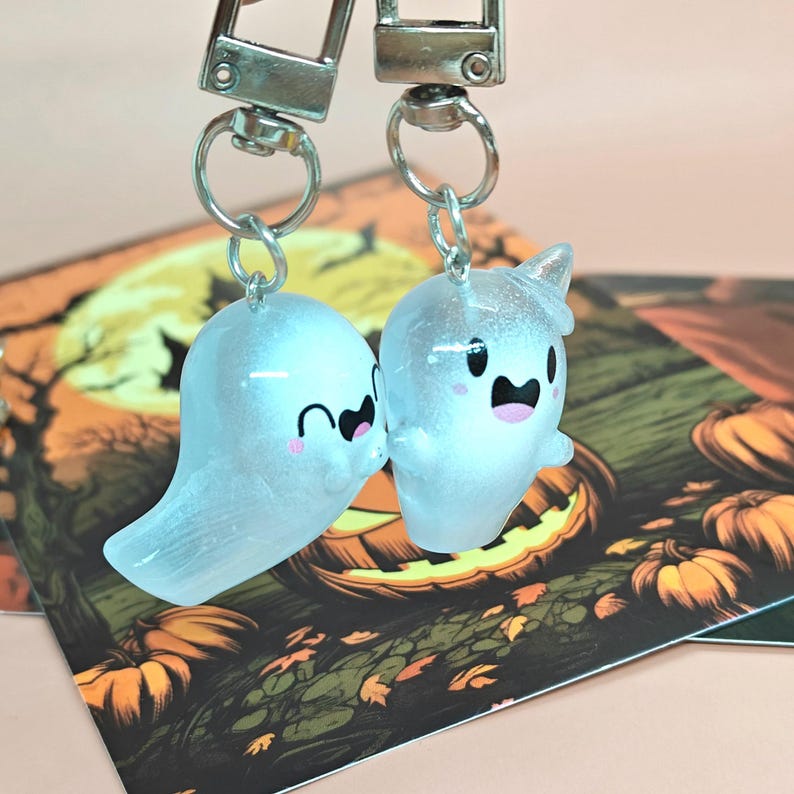 May include: Two clear ghost-shaped keychains with silver hardware. One ghost has a curved smile, the other has a small horn. The background features a Halloween-themed illustration with pumpkins and a full moon.