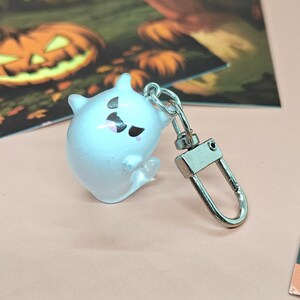 May include: A small, translucent white ghost-shaped keychain with black and pink facial details. The keychain has a silver-colored clasp. The background includes a Halloween-themed illustration.