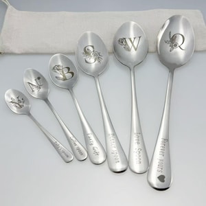 May include: A collection of six stainless steel spoons, each engraved with a floral design and a letter. The spoons are inscribed with phrases like "Baby's spoon" and "Forever yours." They are displayed on a white surface.