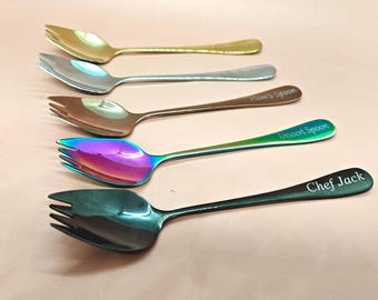 May include: Five spoons with fork-like tines. The spoons are in gold, silver, bronze, iridescent, and black. The black spoon has "Chef Jack" engraved on the handle. The iridescent spoon is green and purple. The bronze spoon has "Mom's Spoon" engraved.
