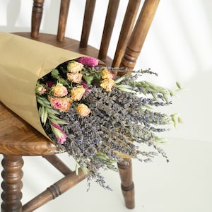 May include: A bouquet of dried flowers wrapped in brown paper, placed on a wooden chair. The bouquet features lavender, dried roses, and other colorful dried flowers. The chair is made of wood and has a traditional design.