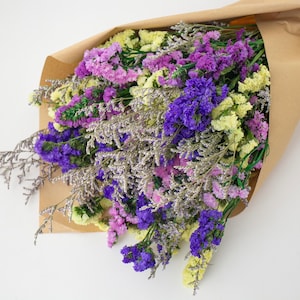 May include: A bouquet of dried flowers wrapped in brown paper. The flowers are a mix of purple, lavender, and yellow hues, with silver-toned accents. The arrangement is presented on a white background.