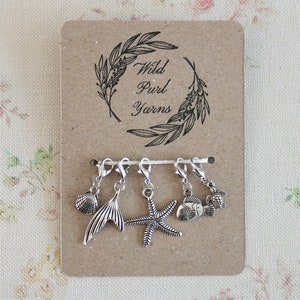 May include: A set of six silver stitch markers with sea-themed charms, including a seashell, a mermaid tail, a starfish, a fish, and a turtle. The stitch markers are displayed on a brown card with the text "Wild Purl Yarns" in a wreath.