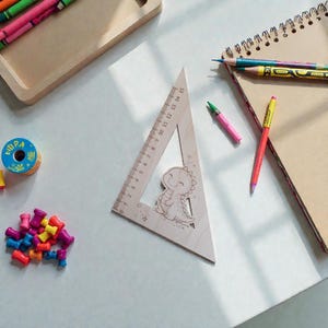 Puede incluir: Una regla triangular de madera con una ilustración de dinosaurio sonriente, que mide hasta 15 pulgadas (aproximadamente 38 cm). La regla está rodeada de lápices de colores, crayones y artículos de papelería, creando un espacio de trabajo creativo.