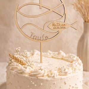 Puede incluir: Una tarta glaseada blanca con un adorno de pastel de madera. El adorno presenta una cruz, un pez y la palabra "Taufe" con el nombre "Matheo" y la fecha 03.05.2026. La tarta está decorada con glaseado blanco y pequeñas perlas blancas.