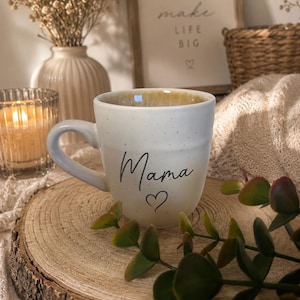 Könnte beinhalten: Ein gesprenkelter, cremeweißer Keramikbecher mit dem Wort Mama in schwarzer Schreibschrift und einem kleinen Herzsymbol darunter. Der Becher hat einen abgerundeten Henkel und eine leicht konische Form mit einer glatten Oberfläche und dunklen Punkten.