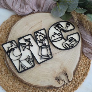 May include: Three camping-themed wooden cutouts spelling "WNC". Each letter features a black outline with camping-related imagery: a tent, campfire, lantern, mountains, and a camper. The cutouts are set on a wooden slice.