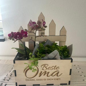May include: A wooden planter box with a decorative picket fence design, holding a vibrant orchid with deep purple blooms and lush green plants. The box features the engraved text "Beste Oma" and "DANKE, DASS ES DICH GIBT!"