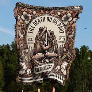 Op de afbeelding: Een geweven wandkleed met een bloemenmotief en twee kikkers die elkaar omhelzen. Het wandkleed heeft de tekst "Till Death Do Us Part" met de data "Est. 2021" en de namen "Ryan & Juliana".