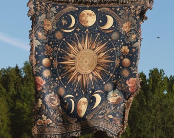 Puede incluir: Una manta de tejido de tapicería con fondo azul oscuro que presenta un diseño de sol y luna dorados con detalles florales.