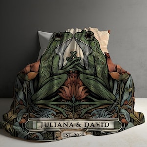 May include: A green and brown throw blanket with a floral design and two frogs facing each other. The blanket has the text "Juliana & David EST 2021" in a banner at the bottom.