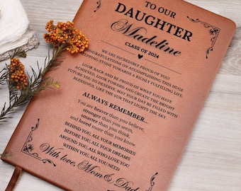 May include: A brown leather journal with a personalized message for a graduate. The journal has a gold border and the text "To Our Daughter Madeline Class of 2024" is written on the front cover. The message inside the journal reads "We are incredibly proud of you! Congratulations on accomplishing this huge stepping stone towards a richly fulfilling life. Look back and be proud of what you have achieved, look forward and create the bright future you so deserve. May your day be filled with blessings, like the sun that lights the sky. Always Remember... You are braver than you believe, stronger than you seem, smarter than you think, and loved more than you know. Behind you, all your memories. Before you, all your dreams. Around you, all who love you. Within you, all you need. With love, Mom & Dad"