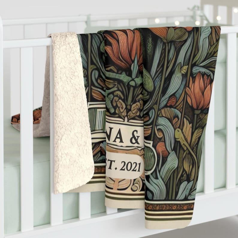 May include: A cream-colored fleece throw blanket with a floral patterned design in shades of green, blue, and orange. The blanket has a decorative border and the text "JA &  EST. 2021" in a vintage font.