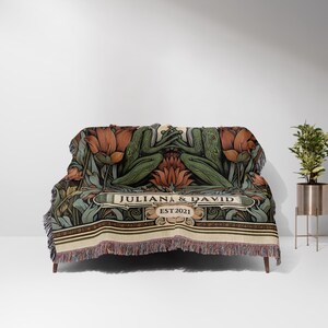 May include: A woven throw blanket with a green and brown floral design featuring two frogs holding hands. The blanket has a cream-colored border with the text "JULIANA & DAVID EST 2021" in a brown banner.