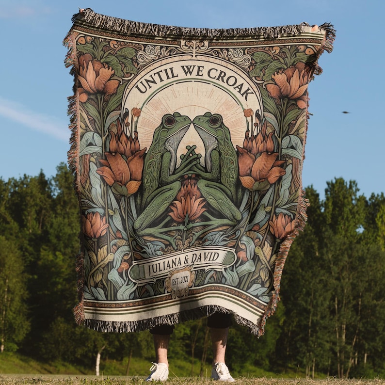 May include: A tapestry with a floral design featuring two green frogs facing each other. The tapestry has the text "Until We Croak" above the frogs and "Juliana & David Est. 2021" below the frogs.