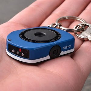 以下が含まれることがあります： 黒い円形レンズとキーリングに付けられた銀色の鍵が付いた、青と白のキーフォブ。キーフォブには番号669701が印刷されています。キーフォブは人の手に持たれています。