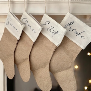 May include: Four beige burlap Christmas stockings with white tops hang from a mantelpiece. Each stocking has a name written in black script: Sienna, Grace, Elliott, and Brooke. The stockings are suspended by a loop of twine.