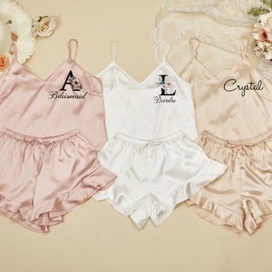 May include: Three sets of matching pajamas with a satin finish. The pajamas are pink, white, and beige. Each set has a tank top with a floral initial and a name, and matching shorts with a ruffle trim. The names are "Bridemaid", "Deirdre", and "Crystal".