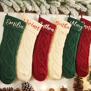 Könnte beinhalten: Sechs personalisierte Weihnachtsstrümpfe mit gestickten Namen auf der Manschette. Die Strümpfe sind aus einem gestrickten Kabelmusterstoff in Grün, Weiß und Rot.