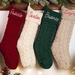 Könnte beinhalten: Vier Zopfstrick-Weihnachtsstrümpfe in Rot, Creme, Grün und Beige hängen an einem Holzbalken. Jeder Strumpf hat einen umgeschlagenen Bund und ist mit einem Namen in einer Kontrastfarbe bestickt: Gracie, Daxton, Brance und Layna.