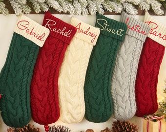 Pode incluir: Seis meias de Natal personalizadas com nomes bordados na bainha. As meias são feitas de tecido de malha de cabo em vermelho, verde, branco e cinza.