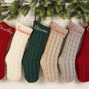 Puede incluir: Seis calcetines de Navidad personalizados con nombres bordados en rojo en el puño. Los calcetines están hechos de una tela gruesa de punto de cable en rojo, blanco, verde, beige y gris.