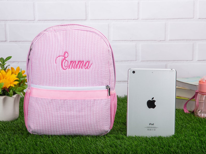 May include: Pink and white striped backpack with a pink zipper and the name "Emma" embroidered in pink on the front.