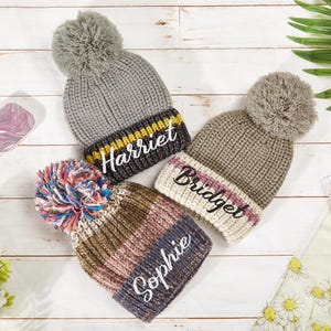 K&ouml;nnte beinhalten: Drei gestrickte Winterm&uuml;tzen mit Bommeln. Die M&uuml;tzen sind grau, braun und rosa mit wei&szlig;en Streifen. Jede M&uuml;tze hat einen Namen bestickt: Harriet, Bridget und Sophie.