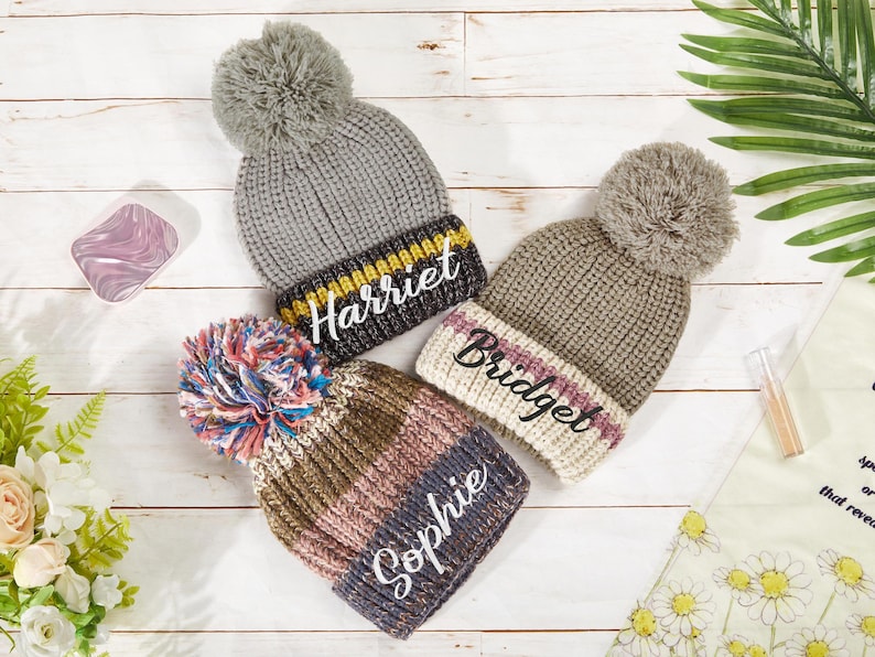 K&ouml;nnte beinhalten: Drei gestrickte Winterm&uuml;tzen mit Bommeln. Die M&uuml;tzen sind grau, braun und rosa mit wei&szlig;en Streifen. Jede M&uuml;tze hat einen Namen bestickt: Harriet, Bridget und Sophie.