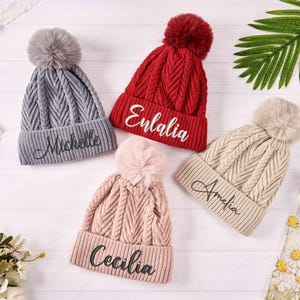 Könnte beinhalten: Vier gestrickte Wintermützen mit Bommeln, jede mit einem anderen Namen auf der Vorderseite bestickt: Michelle, Enlalila, Amelia und Cecilia.