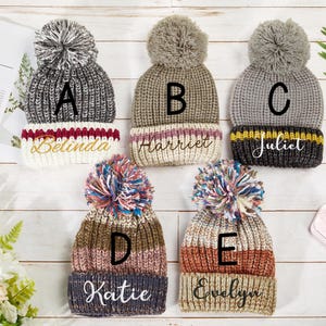 K&ouml;nnte beinhalten: F&uuml;nf gestrickte M&uuml;tzen mit Bommeln in verschiedenen Farben und Mustern. Jede M&uuml;tze hat einen anderen Namen auf dem Umschlag gestickt: Belinda, Harriet, Juliet, Katie und Evelyn.