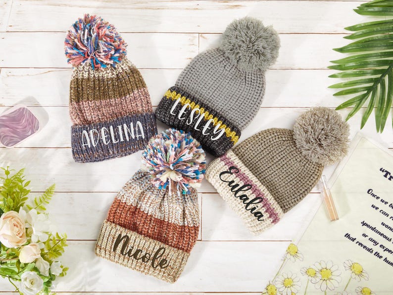 K&ouml;nnte beinhalten: Vier gestrickte Winterm&uuml;tzen mit Bommeln. Die M&uuml;tzen sind in verschiedenen Farben und Mustern gehalten und jede hat einen Namen auf der Vorderseite gestickt: Adelina, Lesley, Eulalia und Nicole.