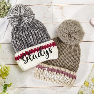 K&ouml;nnte beinhalten: Zwei gestrickte Winterm&uuml;tzen mit Bommeln. Die M&uuml;tze links ist grau und wei&szlig; mit einem roten Streifen und dem Namen "Gladys" bestickt. Die M&uuml;tze rechts ist braun und beige mit einem rosa Streifen und dem Namen "Janice" bestickt.