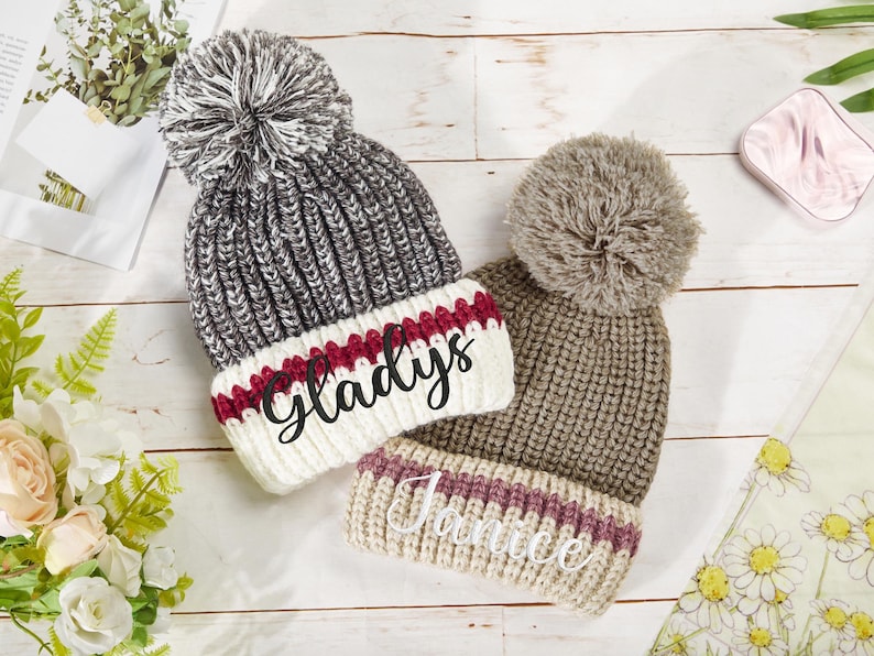 K&ouml;nnte beinhalten: Zwei gestrickte Winterm&uuml;tzen mit Bommeln. Die M&uuml;tze links ist grau und wei&szlig; mit einem roten Streifen und dem Namen "Gladys" bestickt. Die M&uuml;tze rechts ist braun und beige mit einem rosa Streifen und dem Namen "Janice" bestickt.