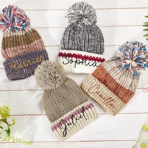 K&ouml;nnte beinhalten: Vier gestrickte Winterm&uuml;tzen mit Bommeln. Die M&uuml;tzen sind in verschiedenen Farben und Mustern gehalten und jede hat einen Namen auf der Vorderseite gestickt: "Harriet", "Sophia", "Camilla" und "Juliet".