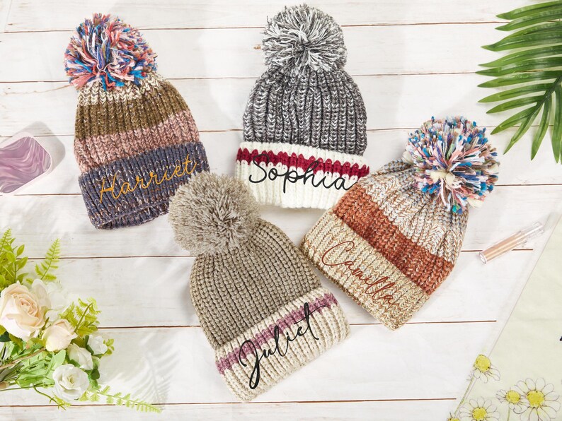 K&ouml;nnte beinhalten: Vier gestrickte Winterm&uuml;tzen mit Bommeln. Die M&uuml;tzen sind in verschiedenen Farben und Mustern gehalten und jede hat einen Namen auf der Vorderseite gestickt: "Harriet", "Sophia", "Camilla" und "Juliet".
