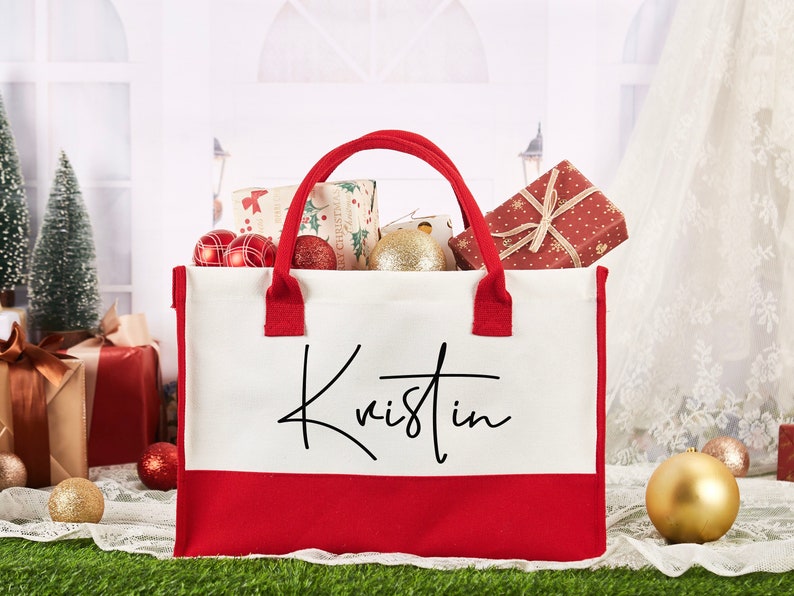 K&ouml;nnte beinhalten: Eine rote und wei&szlig;e Canvas-Tragetasche mit dem Namen "Kristin" in Schwarz bedruckt. Die Tasche ist gef&uuml;llt mit verpackten Geschenken und Weihnachtsornamenten.