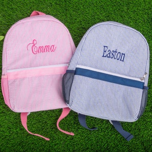 May include: Two personalized striped backpacks, one pink with the name "Emma" embroidered on it and one blue with the name "Easton" embroidered on it.