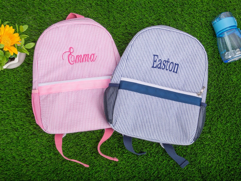 May include: Two personalized striped backpacks, one pink with the name "Emma" embroidered on it and one blue with the name "Easton" embroidered on it.