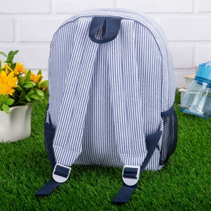 May include: A blue and white striped backpack with navy blue straps and a mesh pocket. The backpack has a top handle and adjustable straps.