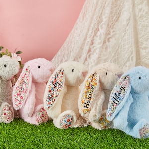 May include: Five plush bunny toys with floral fabric ears. The bunnies are in different colors: gray, pink, cream, beige, and blue. Each bunny has a name embroidered on its ear: Sophia, Lindory, Burly, Heloise, and Hillary.