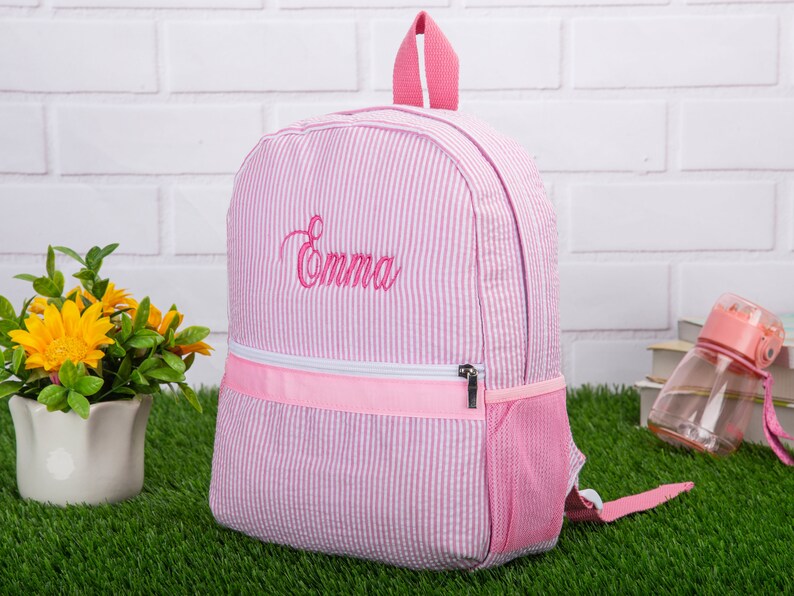 May include: Pink and white striped backpack with a pink zipper and a mesh pocket. The backpack is embroidered with the name "Emma".