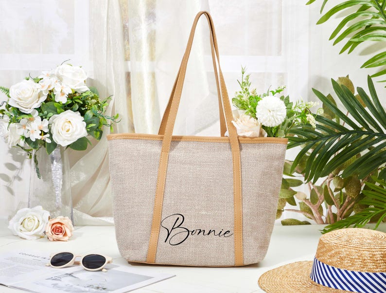 May include: A beige tote bag with tan straps and trim, featuring the name "Bonnie" in black script. The bag is set against a backdrop of flowers, sunglasses, and a straw hat.