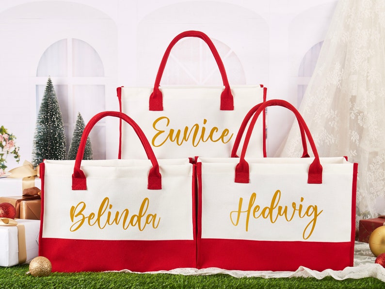 K&ouml;nnte beinhalten: Drei rote und wei&szlig;e Canvas-Tragetaschen mit goldener Beschriftung. Die Taschen sind mit den Namen Eunice, Belinda und Hedwig personalisiert.
