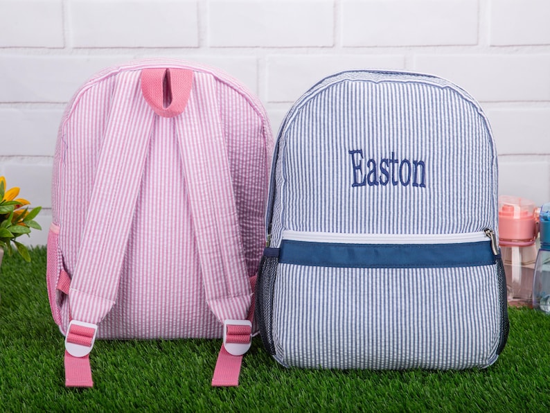 May include: Two striped backpacks, one pink and one blue, with adjustable straps. The blue backpack has the name Easton embroidered on the front.