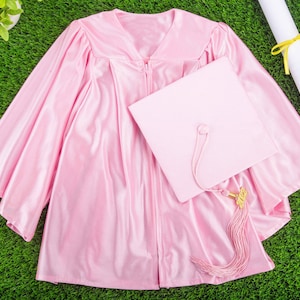 Puede incluir: Toga y birrete de graduación de color rosa con borla dorada. La toga es de satén y tiene cierre de cremallera. El birrete es cuadrado y tiene una borla dorada. La toga y el birrete están sobre una superficie de hierba verde.