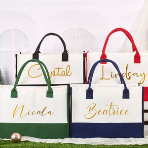 K&ouml;nnte beinhalten: Vier wei&szlig;e Canvas-Tragetaschen mit unterschiedlichfarbigen B&ouml;den und goldenen Schriftz&uuml;gen. Die Taschen sind mit "Nicola", "Beatrice", "Lindsay" und "Crystal" beschriftet.
