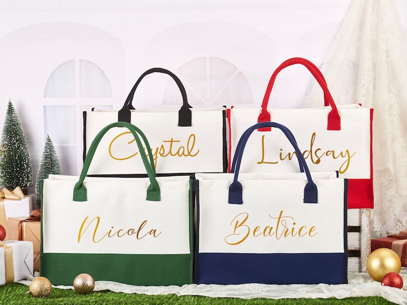 K&ouml;nnte beinhalten: Vier wei&szlig;e Canvas-Tragetaschen mit unterschiedlichfarbigen B&ouml;den und goldenen Schriftz&uuml;gen. Die Taschen sind mit "Nicola", "Beatrice", "Lindsay" und "Crystal" beschriftet.