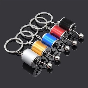 May include: Six keyrings with miniature gear shift designs. Each keyring has a different coloured cylinder: silver, gold, blue, red, and black. The gear shift lever and key ring are silver.